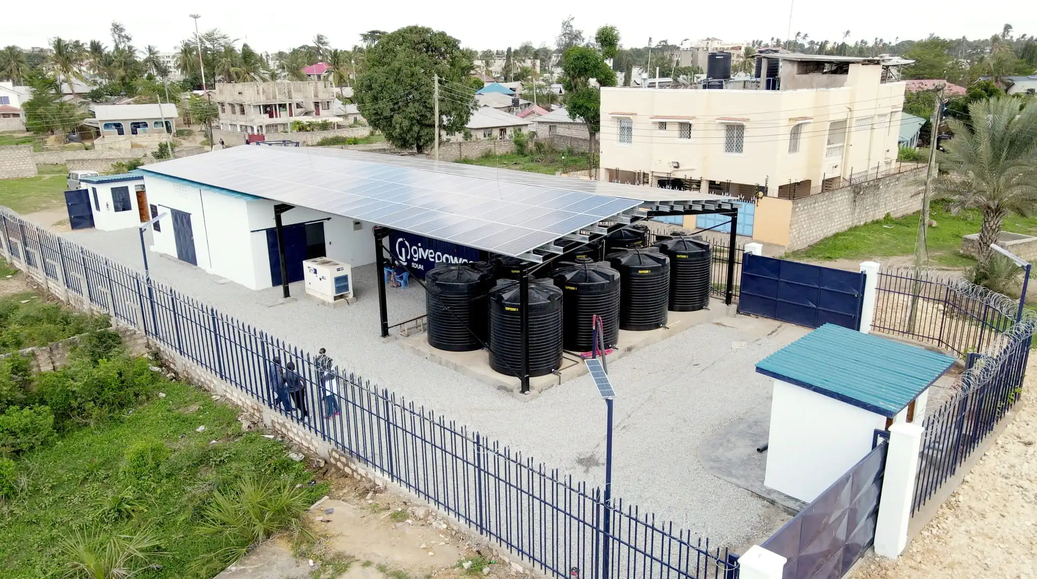 A solar-powered water facility with black tanks, surrounded by a fence, in a residential area.