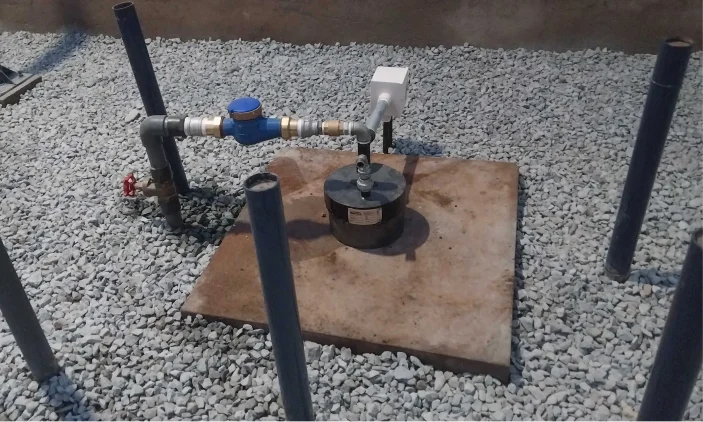 A water meter system with pipes, atop gravel and a concrete slab, with three vertical blue pipes nearby.