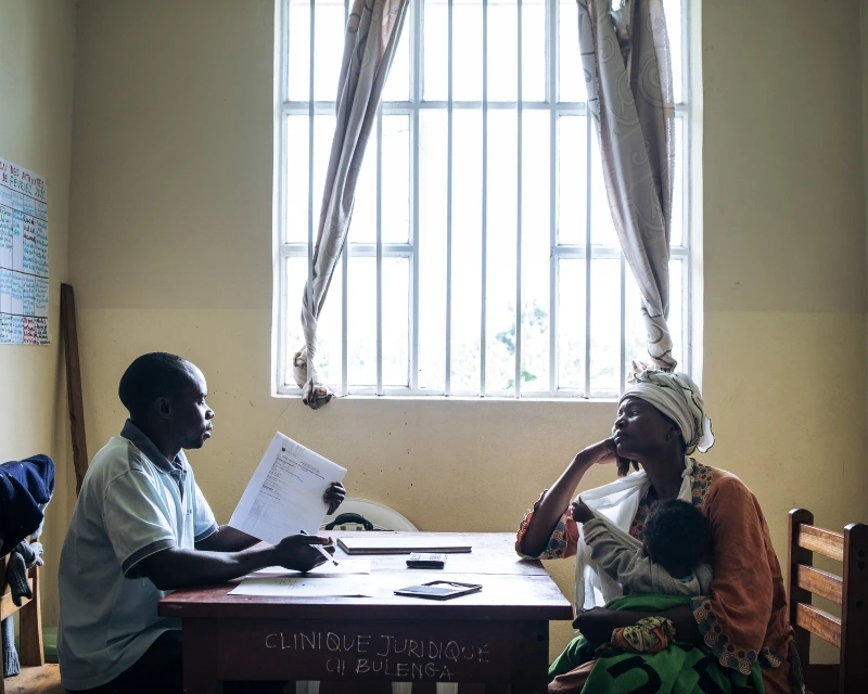 A man and a woman with a child sit across a desk in a sunlit room, discussing documents.