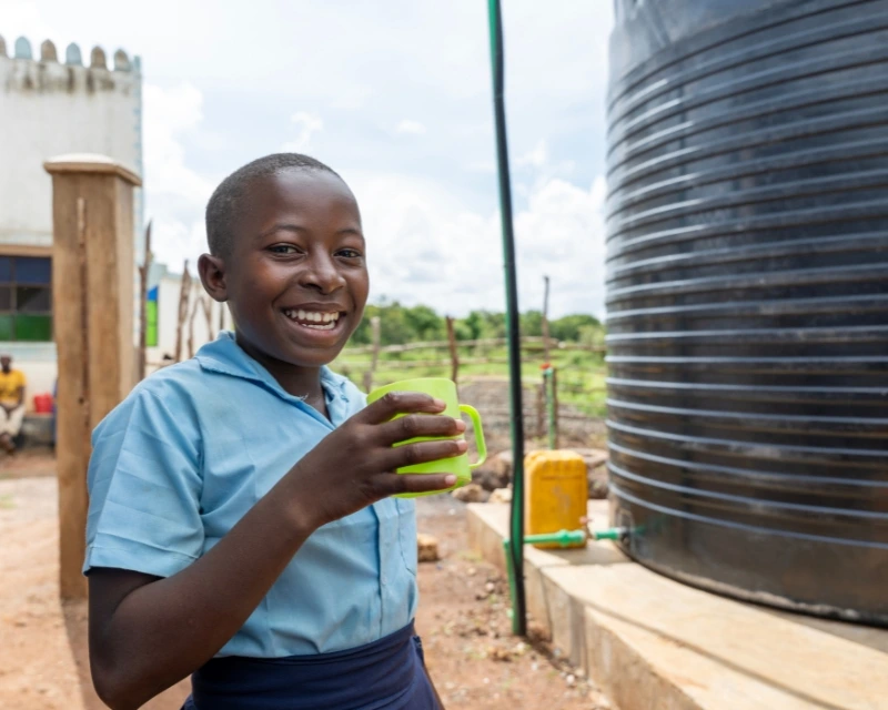 Smiling child in blue shirt holds cup near large black water tank outdoors on a sunny day.