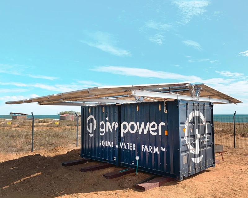 A blue shipping container labeled solar water farm with solar panels on top, set on dry land under a blue sky.