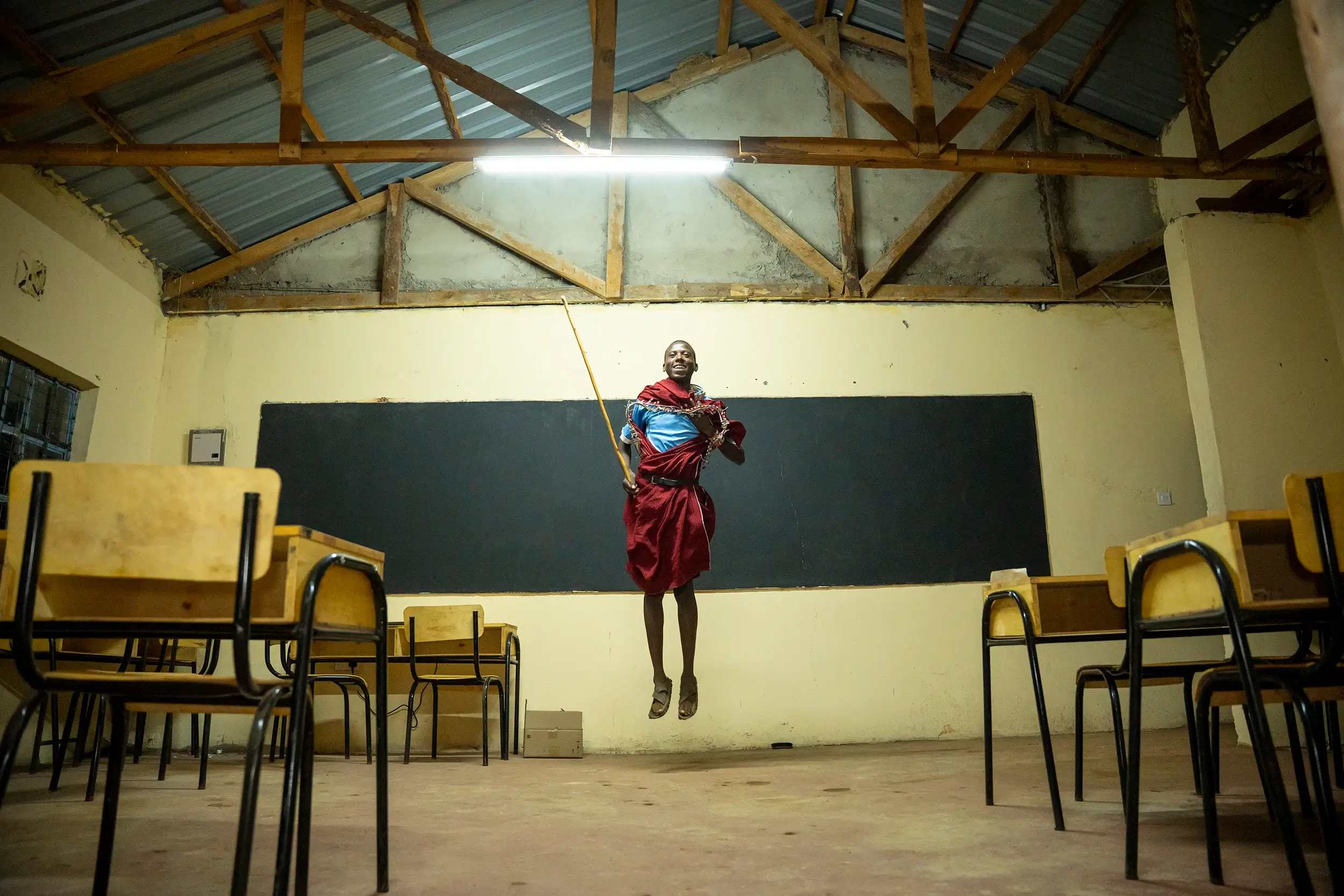 Una persona vestida con ropa tradicional salta en el aire dentro de un salón de clases con pupitres y sillas vacíos.