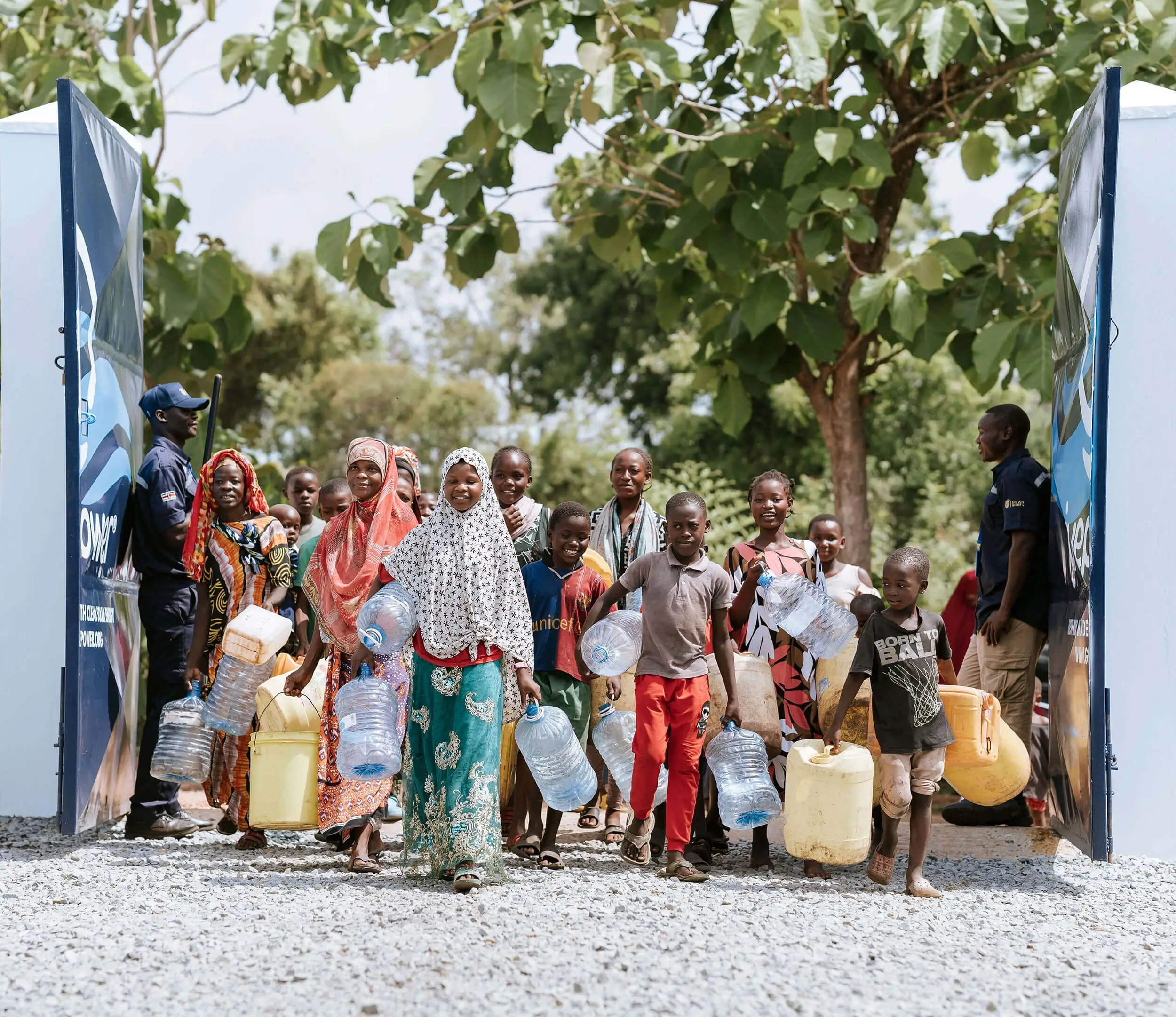 Un grupo de niños y adultos lleva recipientes con agua mientras atraviesa una puerta abierta, con árboles al fondo.