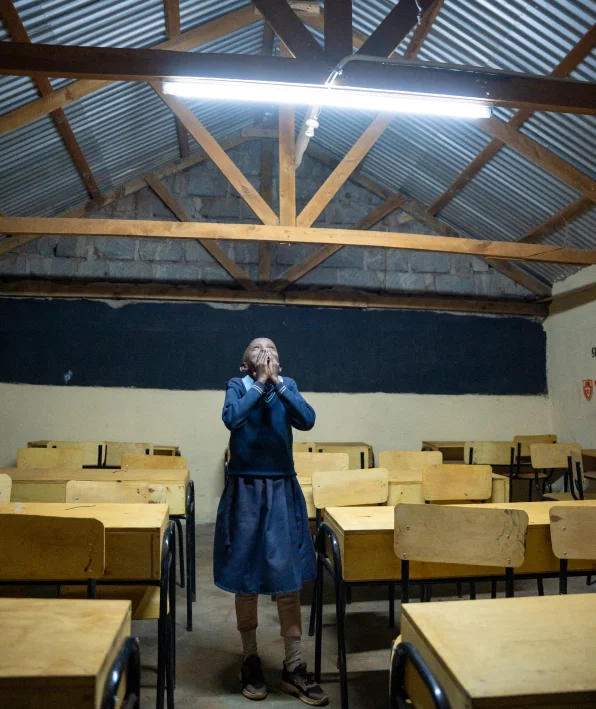 Un estudiante está solo en un salón de clases vacío, con las manos cerca del rostro, bajo las luces fluorescentes.