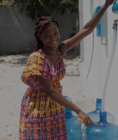 Una mujer con un vestido colorido llena un gran recipiente azul de agua en una estación pública de agua, sonriendo.