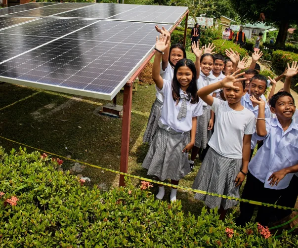 Un grupo de niños en edad escolar saluda con la mano y sonríe junto a unos paneles solares en un entorno al aire libre, frondoso y verde.