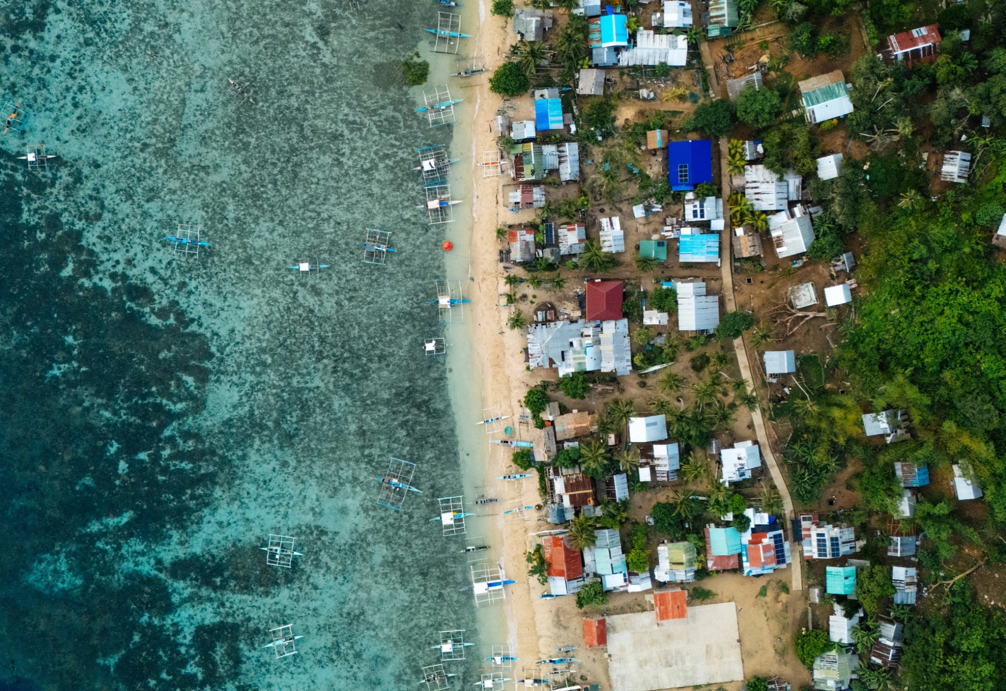 Aerial view of a coastal village with boats along the shoreline and dense buildings next to green vegetation.