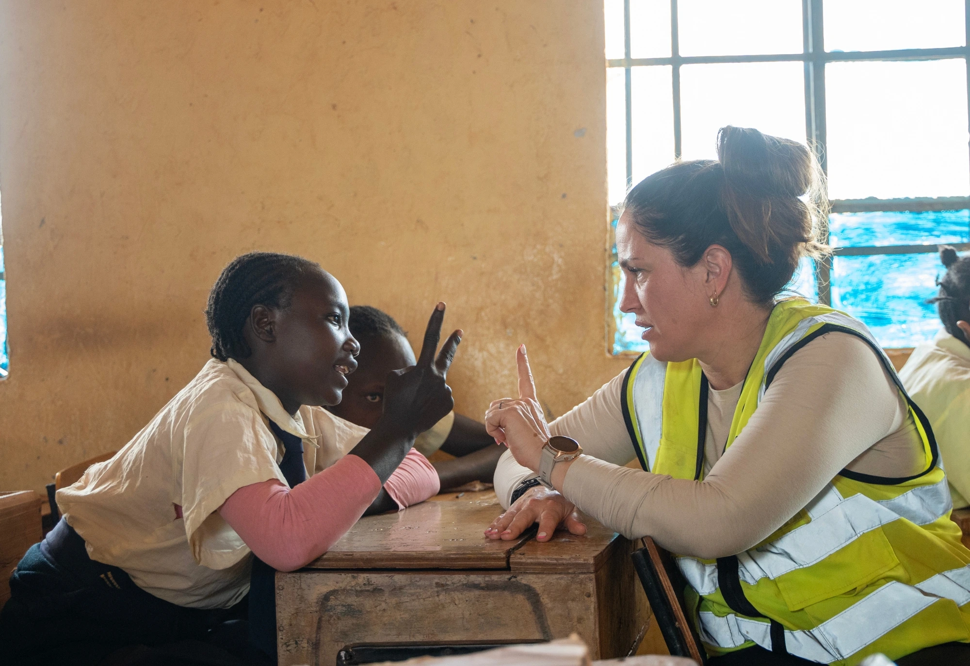 A child and a woman in a safety vest sit at a desk, raising fingers while talking in a classroom.