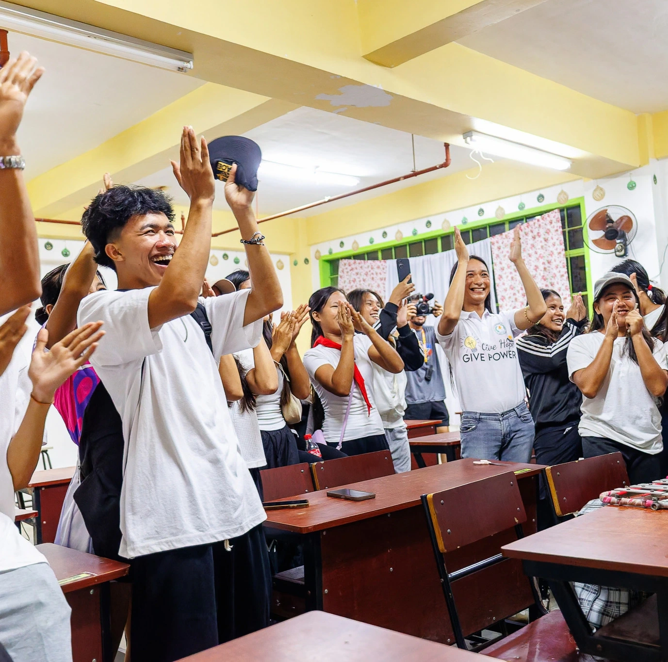 A group of students in a classroom clap, smile, and cheer enthusiastically, standing beside their desks.