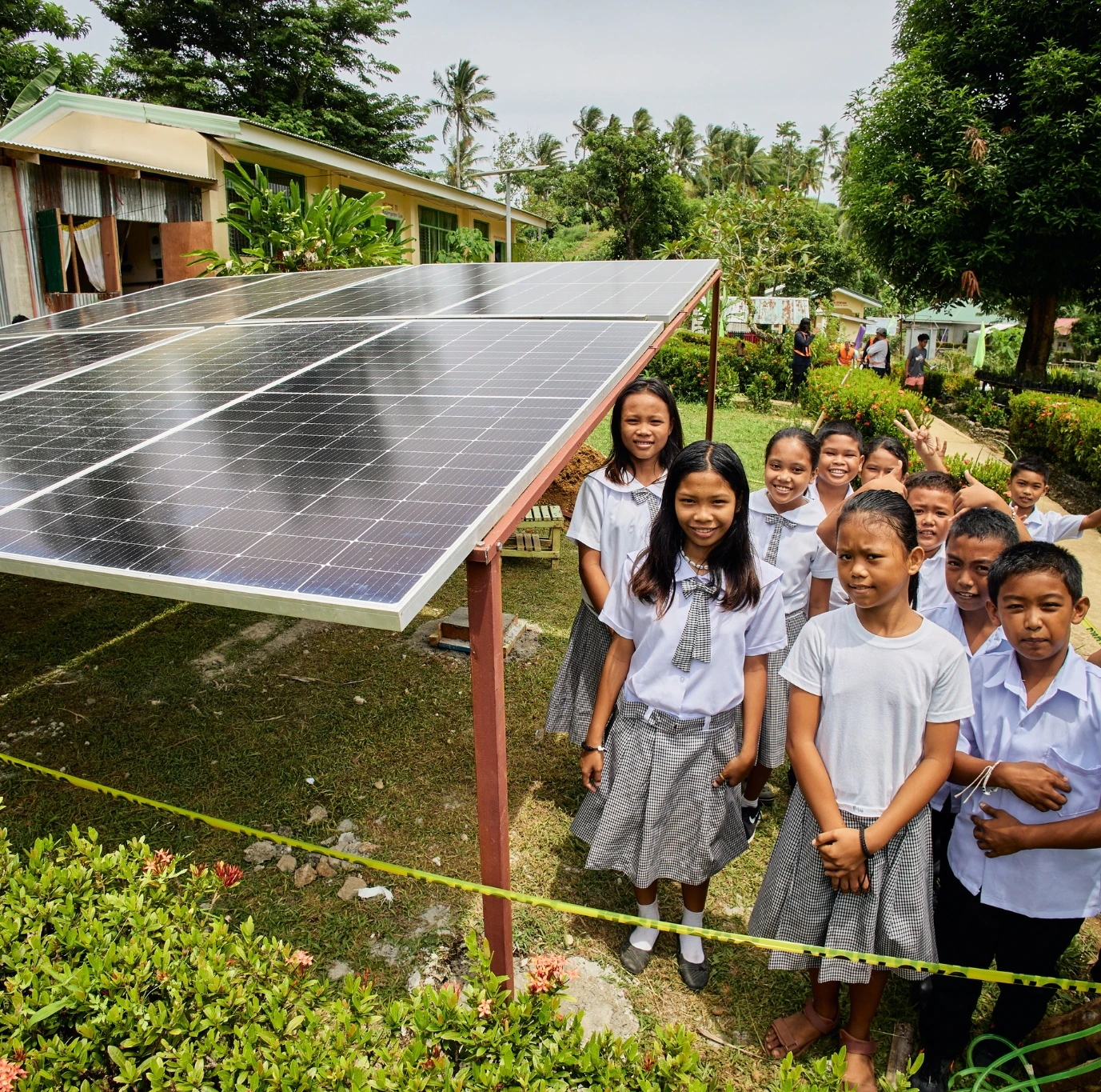 Schoolchildren in uniforms stand by outdoor solar panels in a lush, green garden area, smiling at the camera.