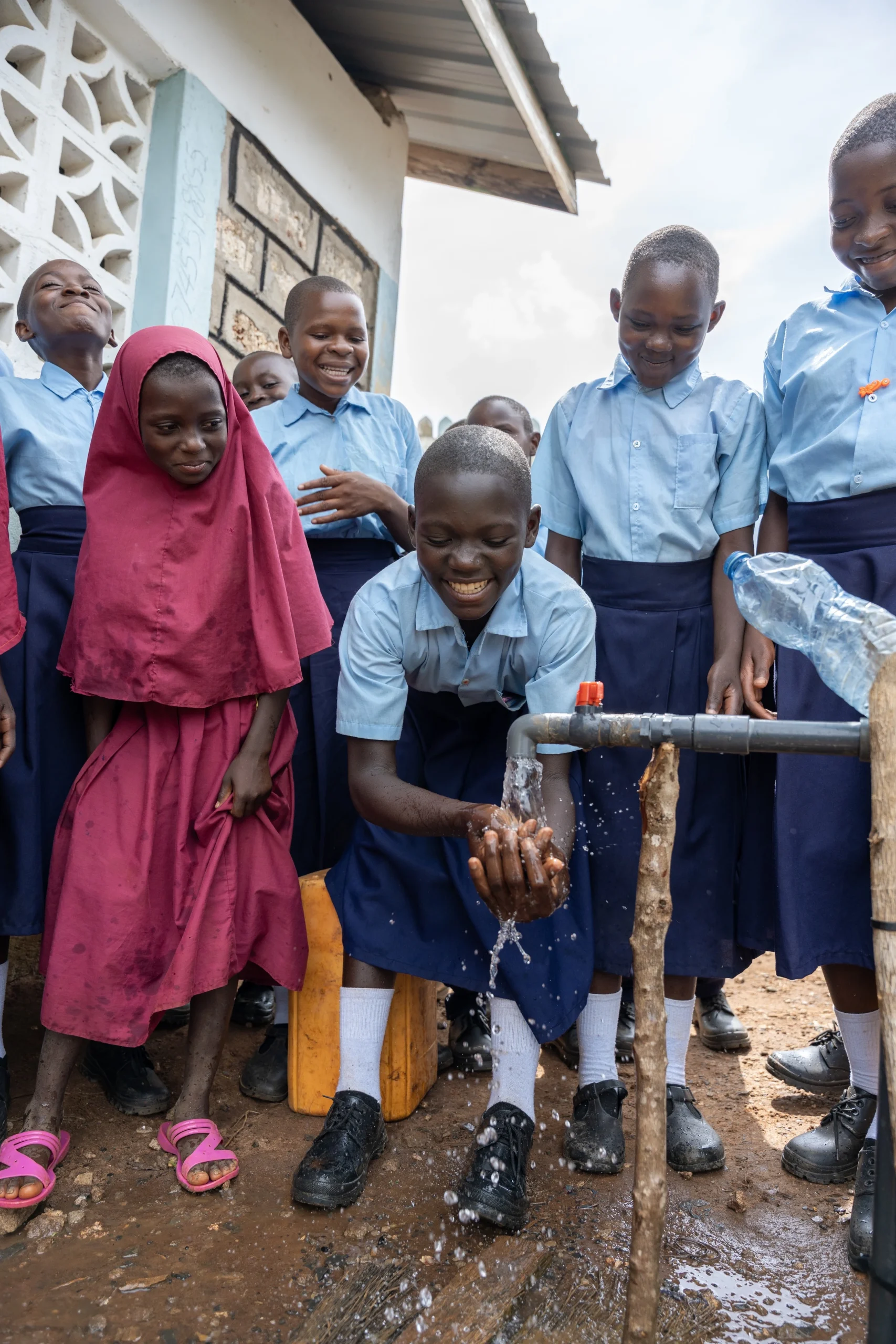 Schoolchildren in uniforms smile as a boy washes his hands at an outdoor water tap.