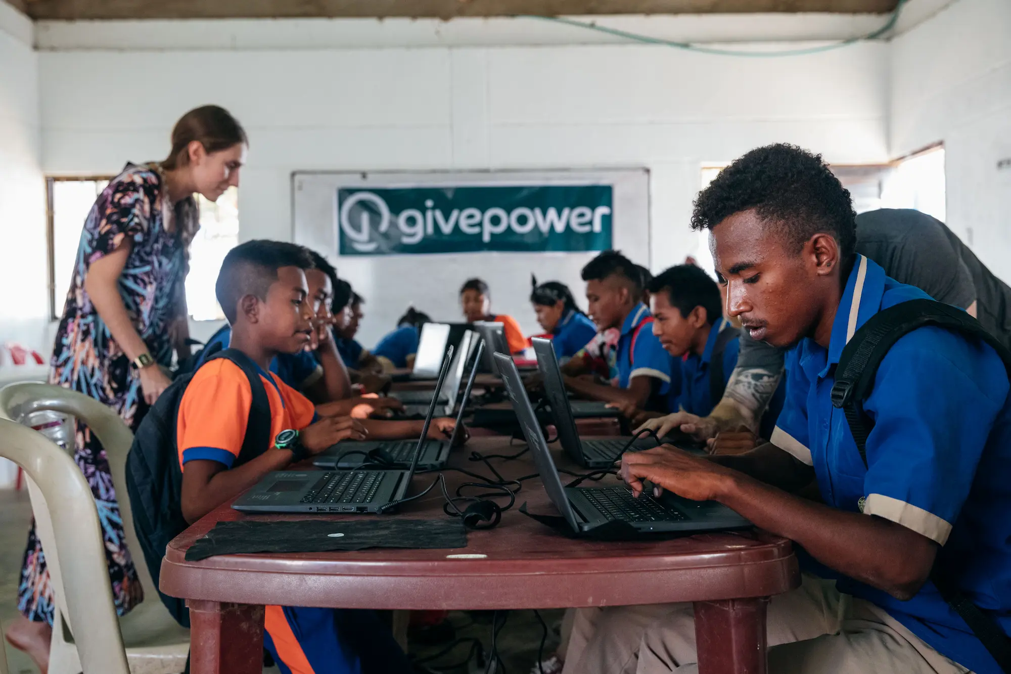 Unos estudiantes con uniformes azules usan computadoras portátiles en una mesa, con un maestro cerca y un cartel de GivePower al fondo.