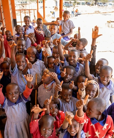 A large group of smiling children in uniforms raise their thumbs up outside a building.