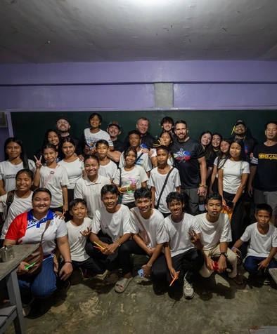 A large group of smiling people, mostly teens and adults, pose together indoors in a brightly lit classroom.
