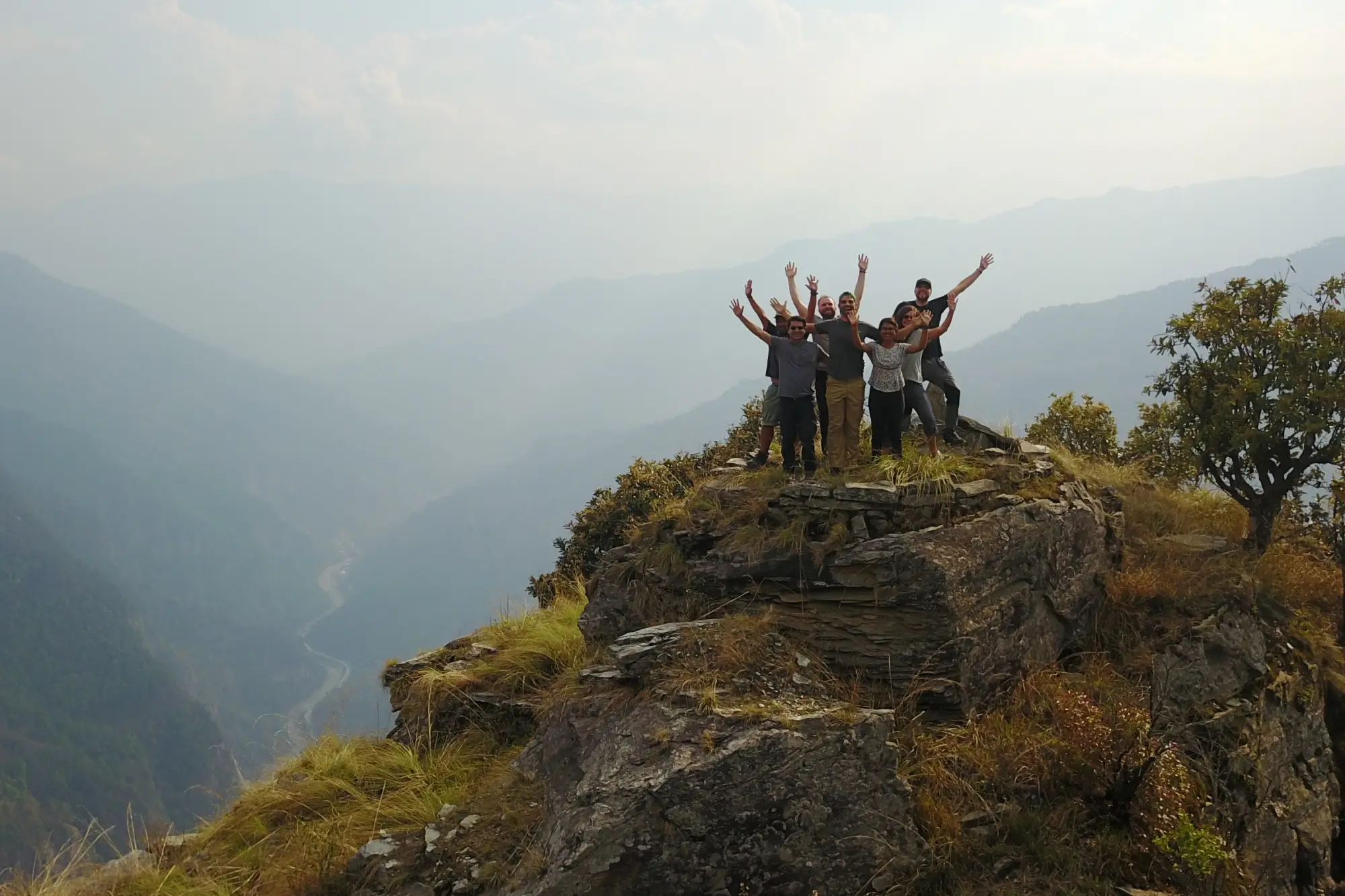 Cinco personas están de pie con los brazos en alto en la cima de una colina rocosa, contemplando un paisaje montañoso y un valle envueltos en la bruma.