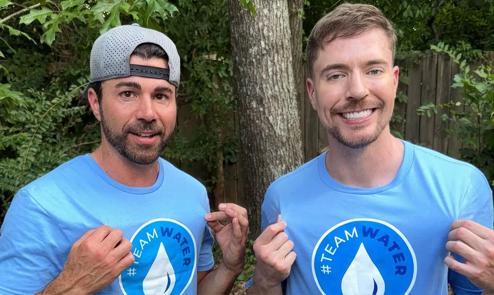 Dos hombres al aire libre, con camisetas azul claro de #TEAM WATER con un logotipo de una gota de agua, de pie junto a un árbol.