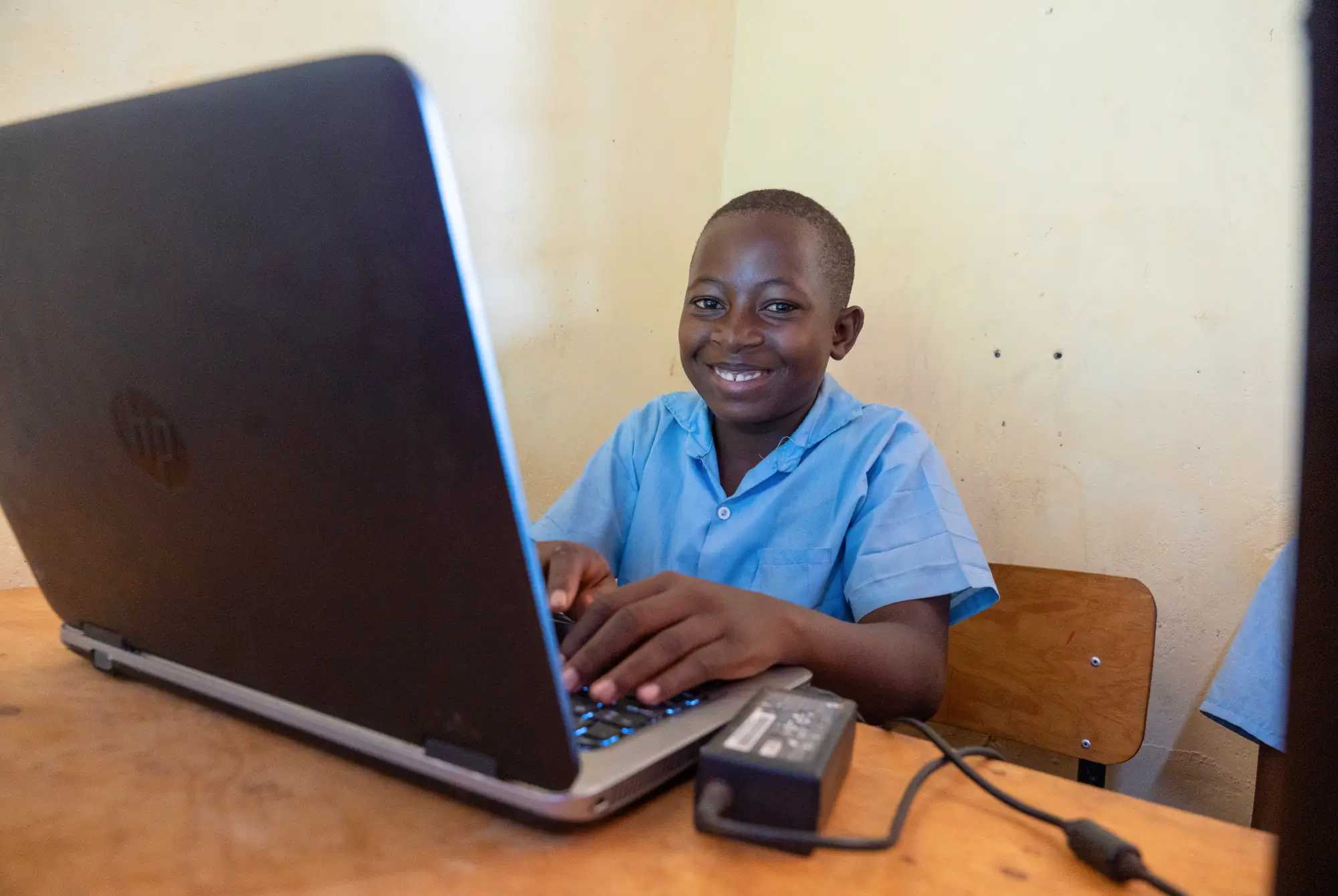 Un niño sonriente con una camisa azul usa una computadora portátil en un pupitre de madera dentro de un salón de clases.