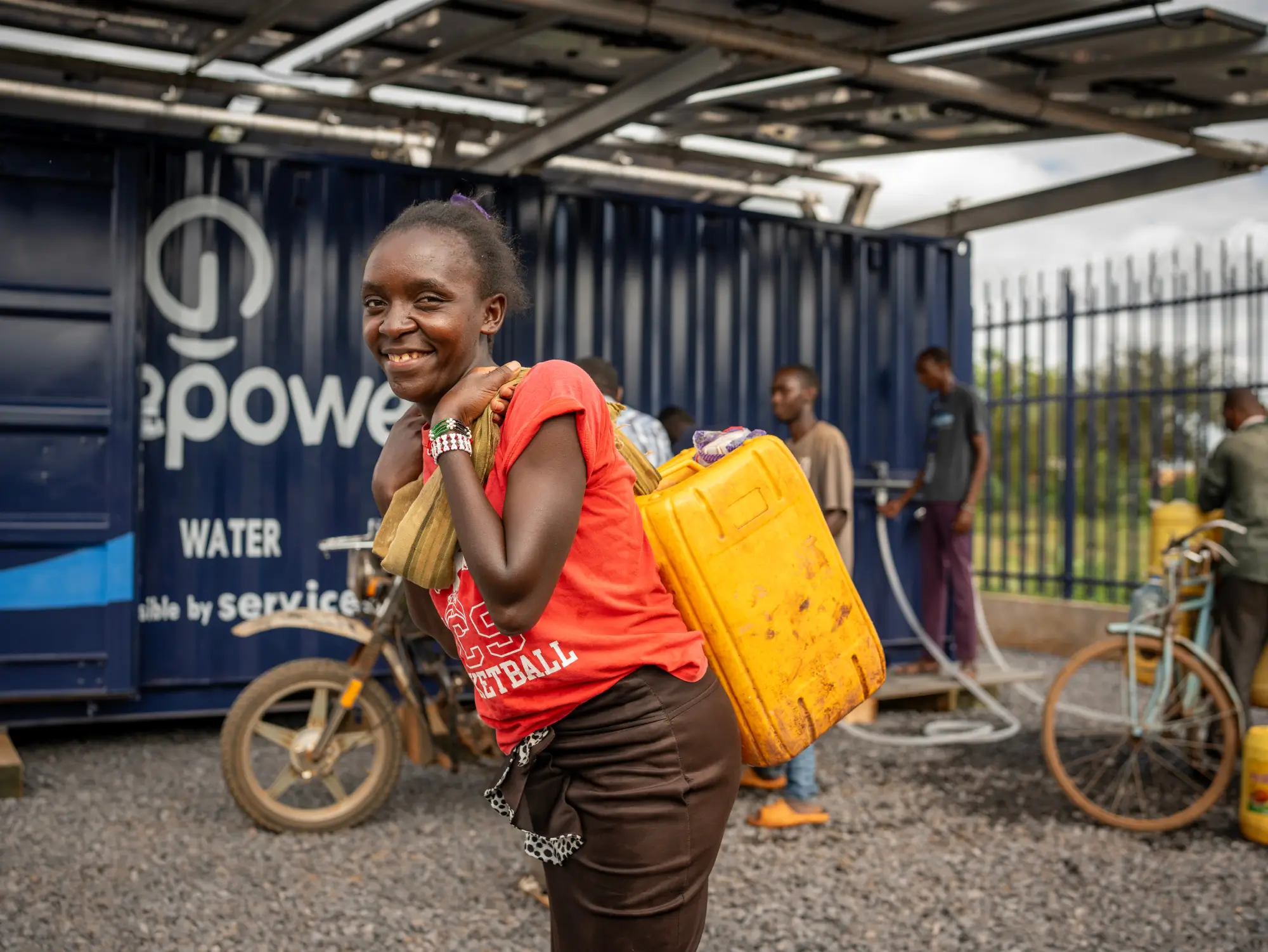 Una mujer sonriente lleva un recipiente amarillo para agua junto a una estación de agua azul, con personas y bicicletas al fondo.
