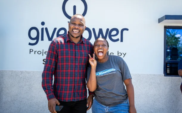 Two people smiling and posing in front of a GivePower sign, one holding up a peace sign.