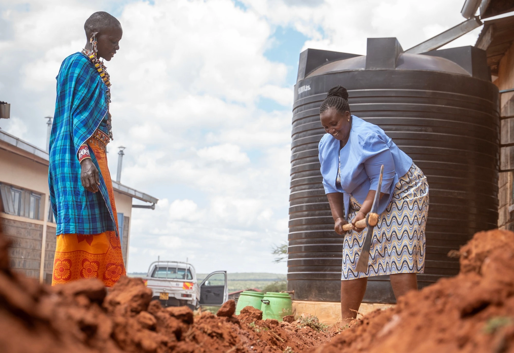 Two women work outside near a large water tank, with soil and buildings visible under a partly cloudy sky.