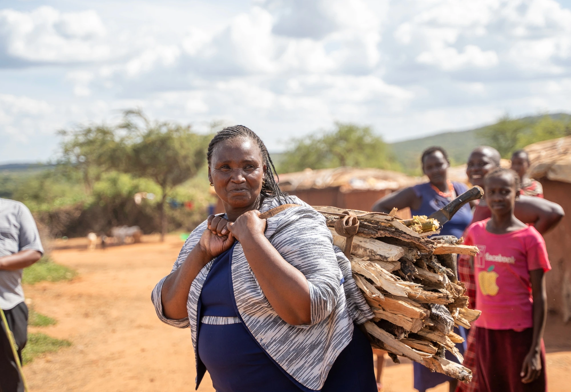 Woman carrying firewood on her back, with people and huts in the background on a sunny day.