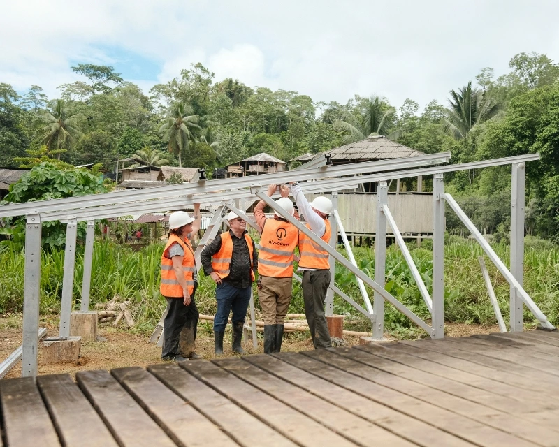 Four workers in orange vests and helmets assemble a metal structure outdoors near wooden houses and greenery.