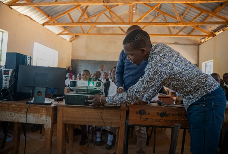 A man adjusts equipment on a desk in a classroom while students watch in the background.