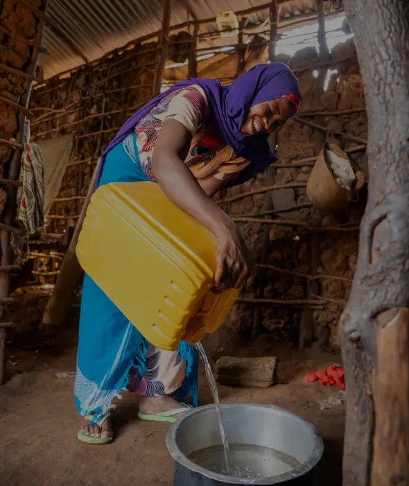 Una mujer vestida con ropa colorida vierte agua de un recipiente amarillo en una olla dentro de un refugio rústico.