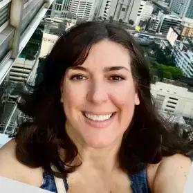 Woman with dark hair smiles, taking a selfie on a rooftop with a cityscape background.