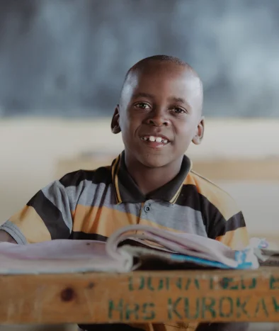 Un niño sonriente con una camisa a rayas está sentado en un pupitre con un libro abierto en un salón de clases.