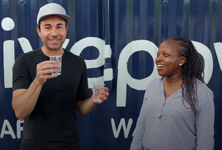 Man and woman smiling, each holding a glass of water, standing in front of a blue wall with white text.