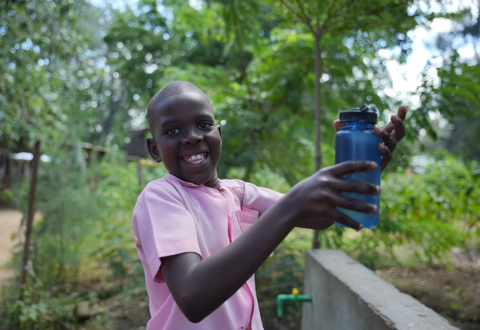 Smiling child in a pink shirt holds a blue water bottle outdoors, surrounded by green trees and plants.