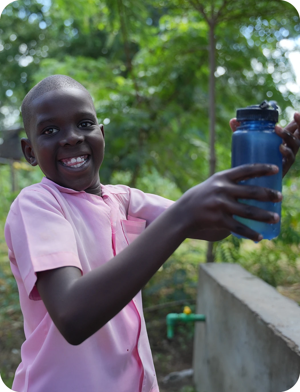 Smiling child in a pink shirt holds a blue water bottle outdoors, with trees and a wall in the background.