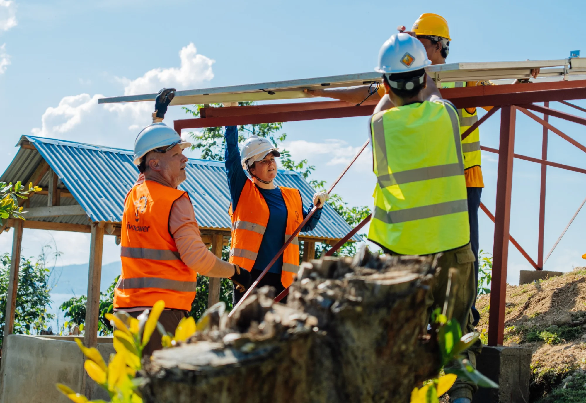 Four people in safety gear work together to build a metal structure outdoors on a sunny day.