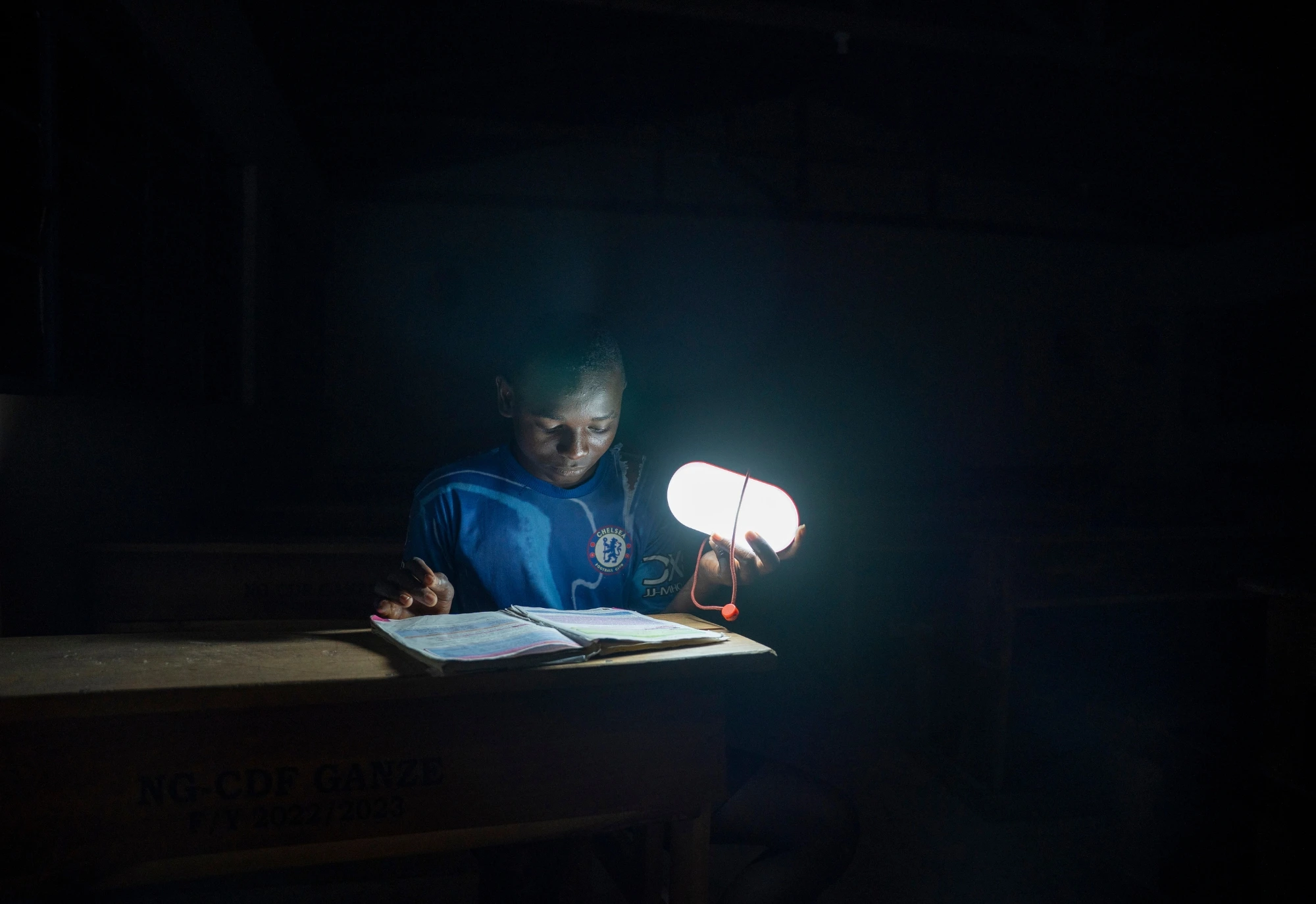 A boy reads a book at a desk, illuminated by a small portable lamp in a dark room.