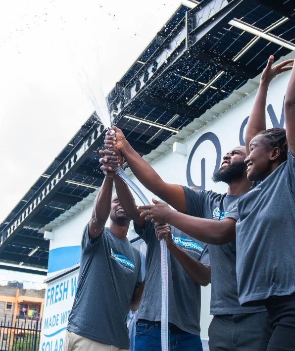 Tres personas rocían agua con una manguera afuera, bajo un techo de paneles solares, sonriendo y estirando los brazos hacia arriba.