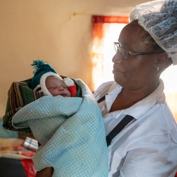 A nurse wearing glasses holds a newborn baby wrapped in a blanket, both indoors with warm lighting.
