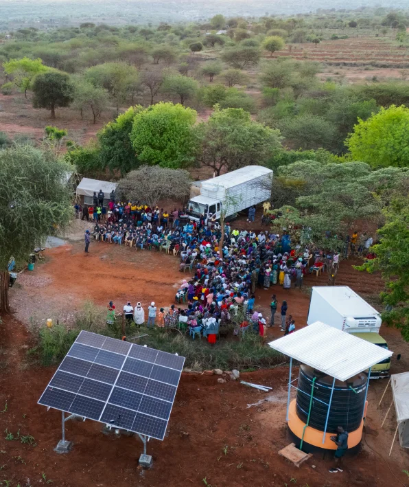 A crowd gathers around trucks in a rural area with trees, near solar panels and a water tank.