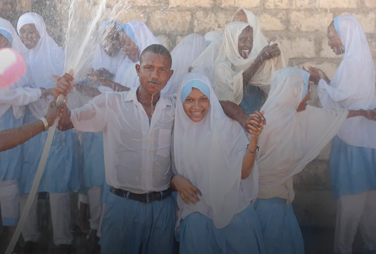 Unos estudiantes sonrientes, vestidos con uniforme, juegan con agua, algunos tomados de la mano, frente a un muro de piedra.