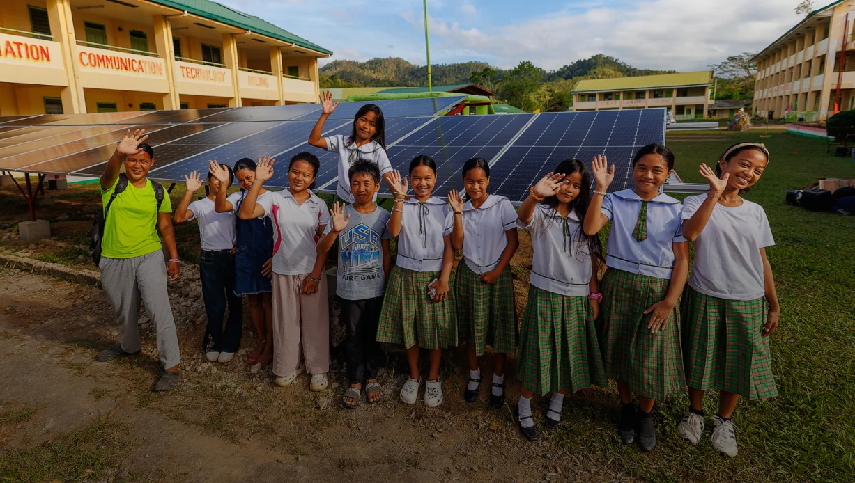 Un grupo de estudiantes sonrientes, vestidos con uniforme, saludan con la mano frente a los paneles solares destinados a escuelas de comunidades remotas.