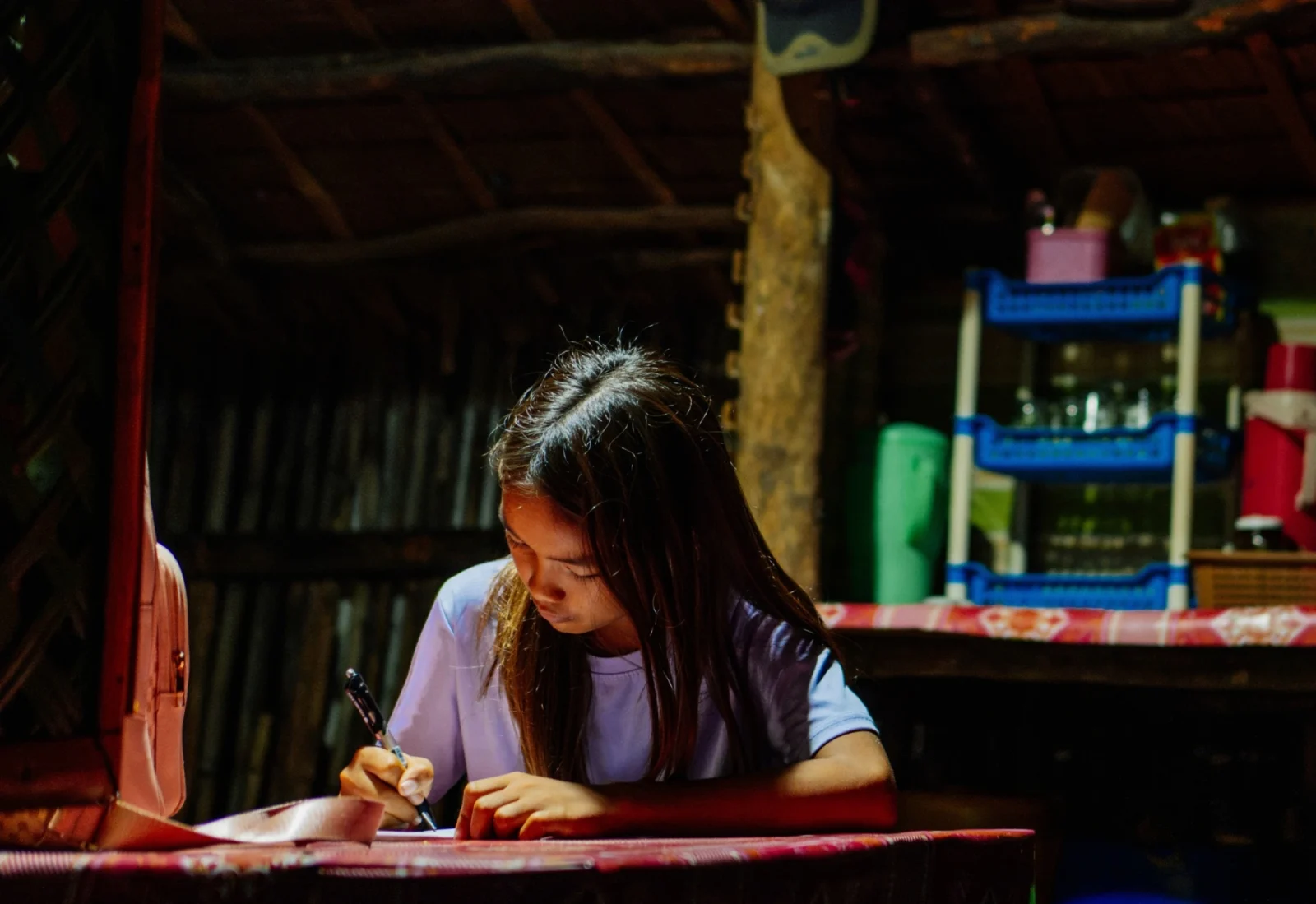 Girl with long hair writing at a table inside a dimly lit rustic kitchen or dining area.