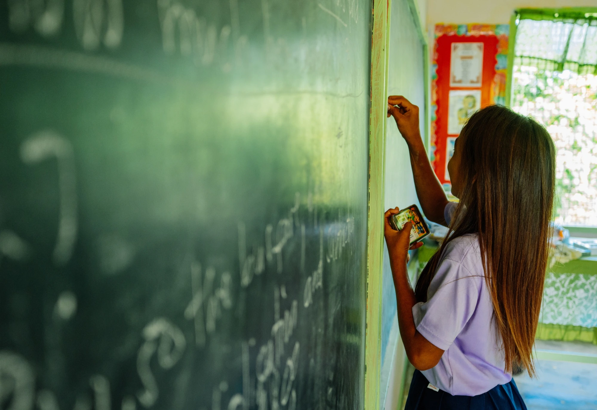 Student with long hair writes on a classroom chalkboard, holding chalk and an eraser in her hands.