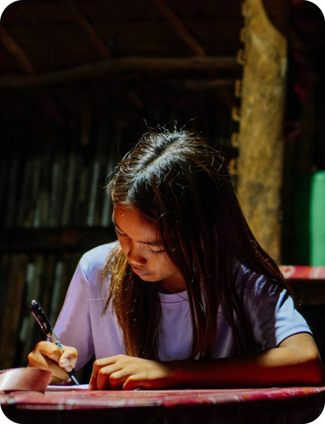 Girl with long hair in a purple shirt writing at a table in a dimly lit room with wooden walls.