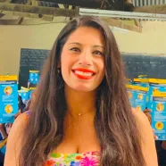 Woman with long dark hair and a bright smile stands in front of colorful packages on shelves.