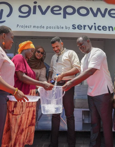 Five people fill a clear container with water at a GivePower water station, smiling and interacting.