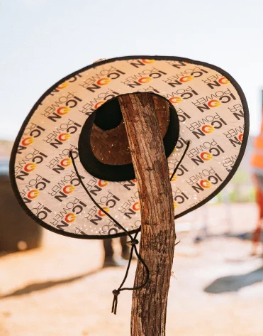 A wide-brimmed hat with ICON print hangs on a vertical tree branch outdoors in sunlight.