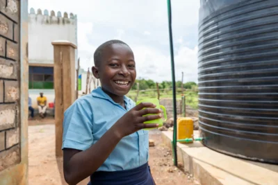 Un niño sonriente, con uniforme escolar, sostiene un vaso verde junto a un gran tanque de agua al aire libre en un día soleado.