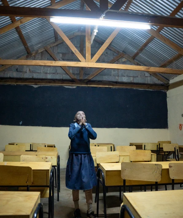 Yon elèv ki gen inifòm ble kanpe poukont li nan yon sal klas vid ki fèb limen, ak biwo an bwa.