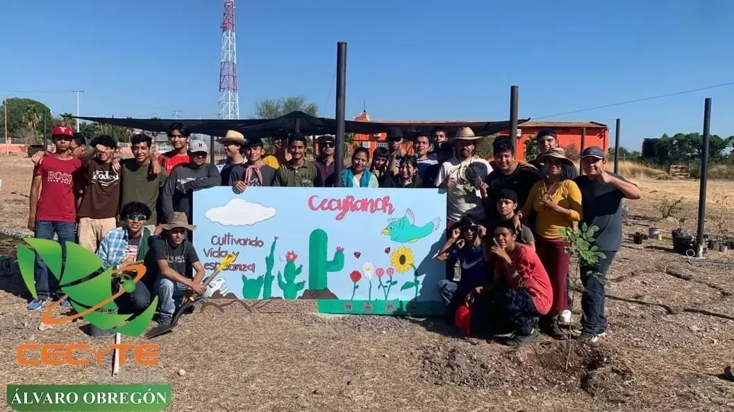 A group of people pose outdoors with a colorful sign that reads Coyaranch Cultivando Vida y Esperanza..