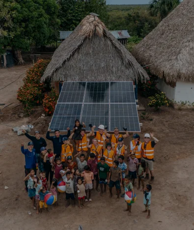 A group of adults and children stand in front of a thatched hut with solar panels, waving at the camera.